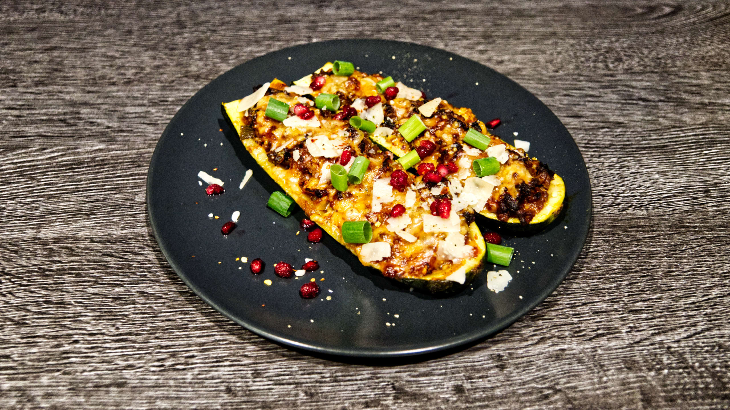 Mit Hackfleisch gefüllte Zucchini, garniert mit Parmesanhobeln, Granatapfelkernen und grünen Frühlingszwiebelringen.