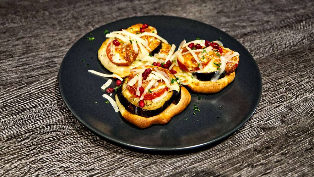 Mini Pizza belegt mit Aubergine und Tomate und mit frisch geriebenem Käse bestreut. Als Farbklecks ein paar Granatapfelkerne.