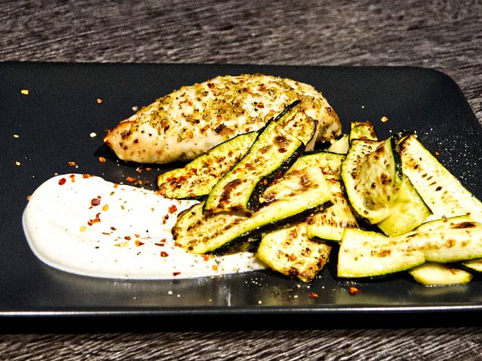 Gebratene Hähnchenbrust an gebratenen Zucchini auf einem schwarzen Teller mit weißem Dip im Vordergrund