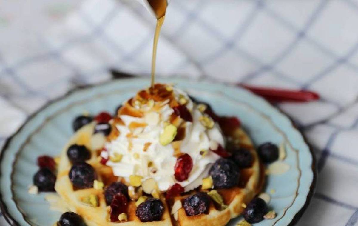 Ahornsirup wird auf eine Waffel mit Blaubeeren und Sahne geschüttet.