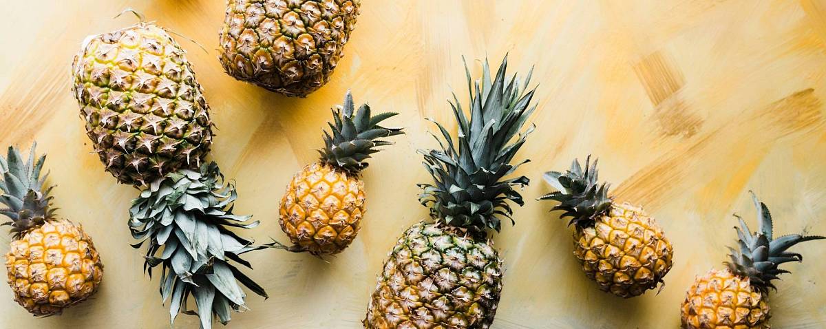 Viele ganze Ananas liegen kreuz und quer auf einem gelben Hintergrund.