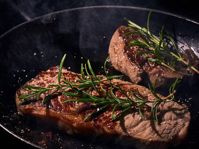 Zwei gebratene Steaks mit Rosmarinzweigen darauf in einer schwarzen Pfanne.