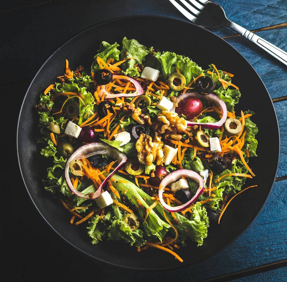 Ein knackiger bunter Salat mit Karottenstiften, Zwiebeln, Oliven und Feta in einem tiefen schwarzen Teller, daneben liegt eine silberne Gabel.